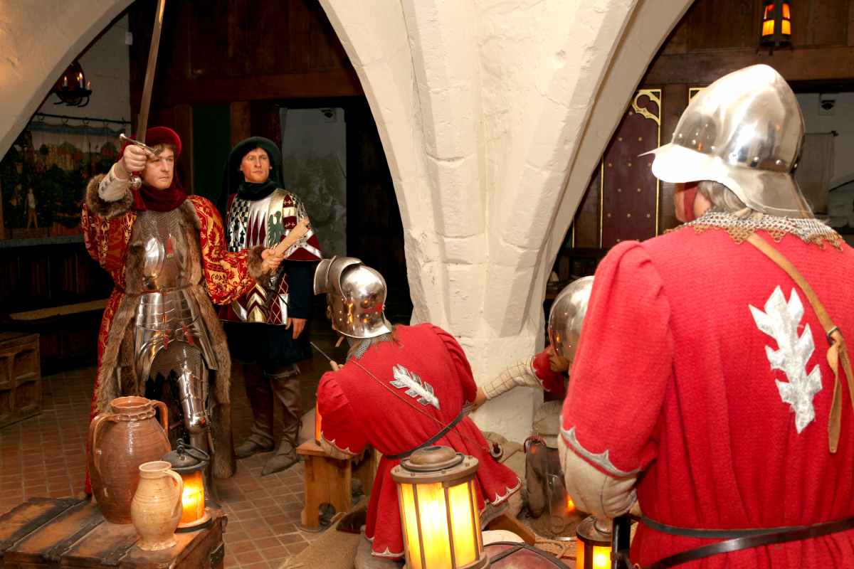 Figures dressed in medieval-style armor and tunics stand in a stone-arched room with lanterns.