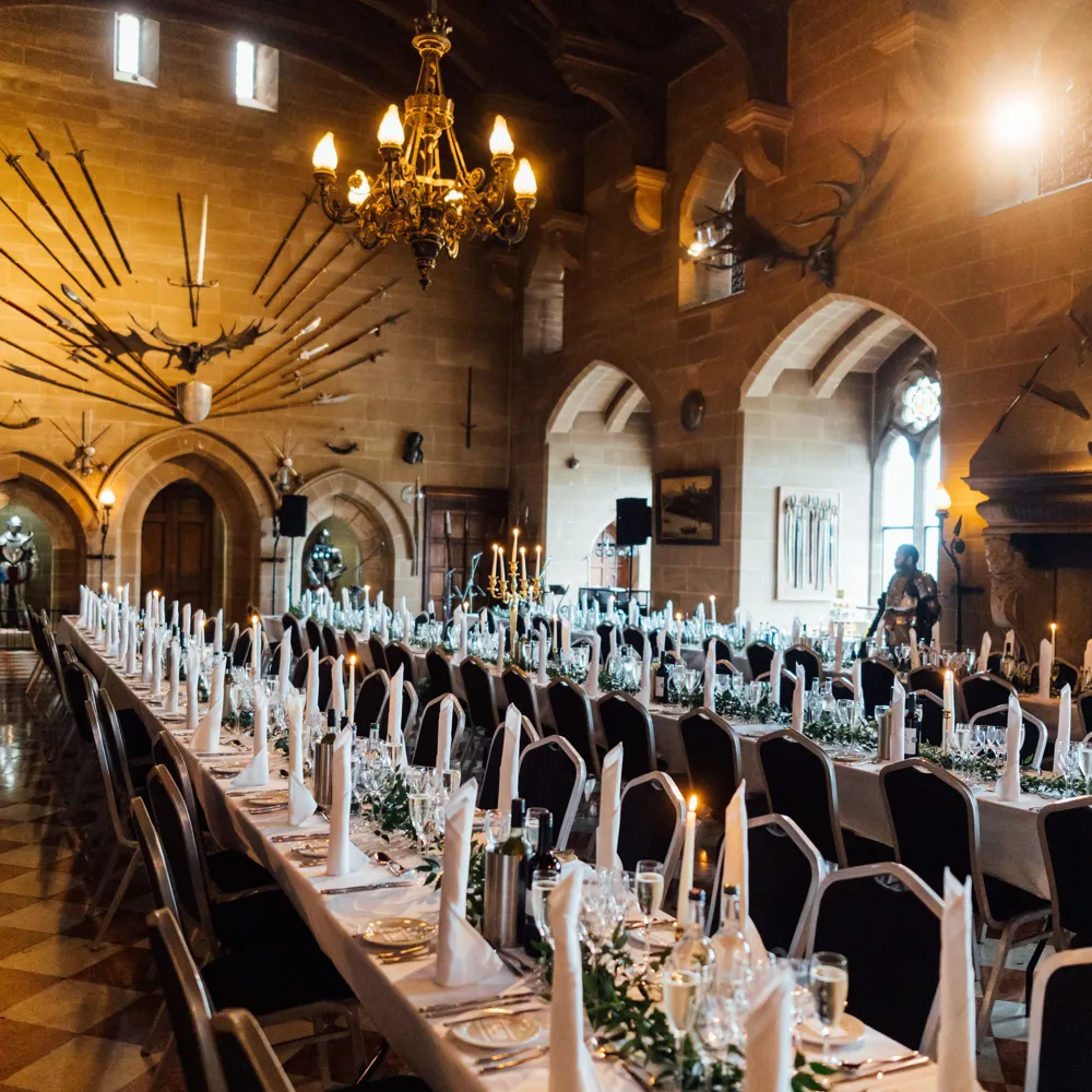 A long banquet table is set for a formal event in a stone hall decorated with wall-mounted swords and a chandelier.