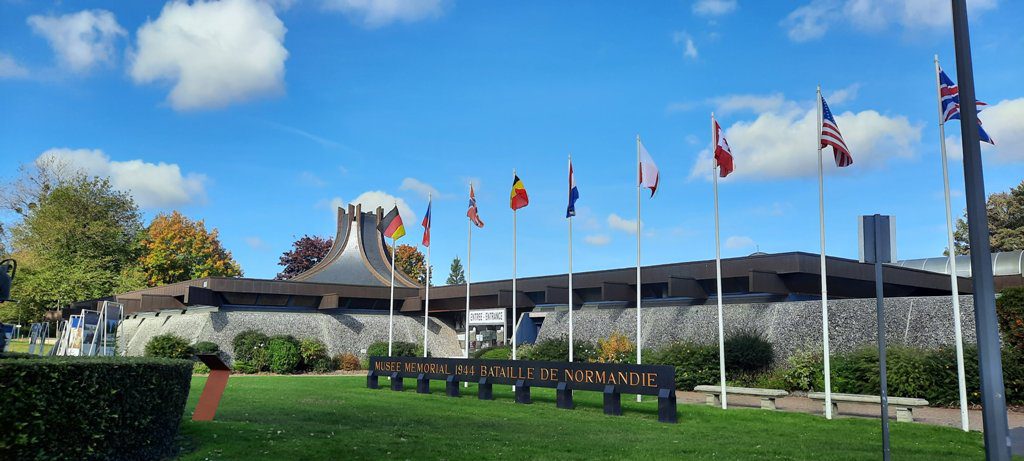 A row of flags stands in front of a building labeled Musee Memorial 1944 Bataille de Normandie under a blue sky.