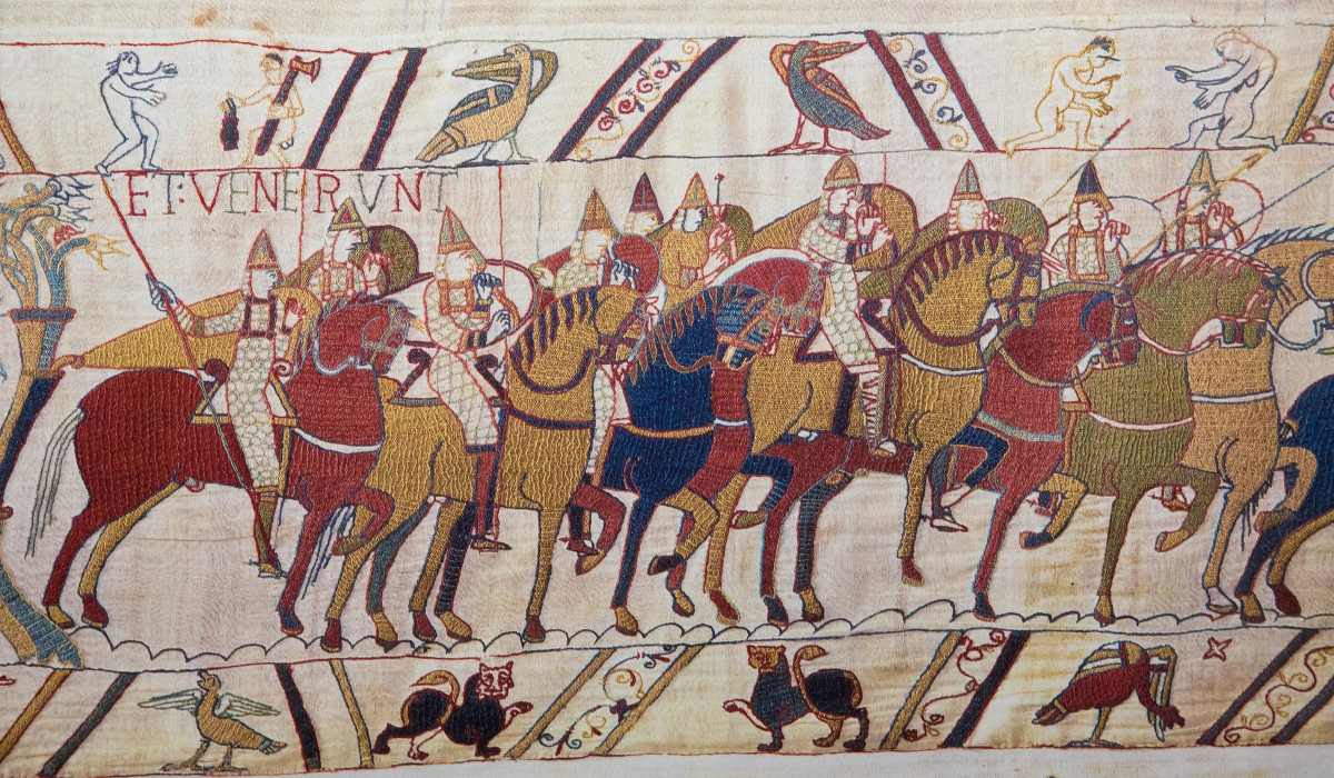 An embroidered tapestry depicts a line of mounted soldiers in conical helmets with Latin text above and animals below.