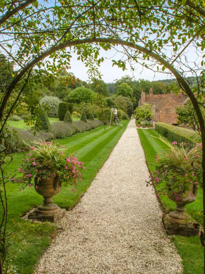 A gravel path leads through a garden framed by an archway, with stone urns filled with flowers in the foreground.