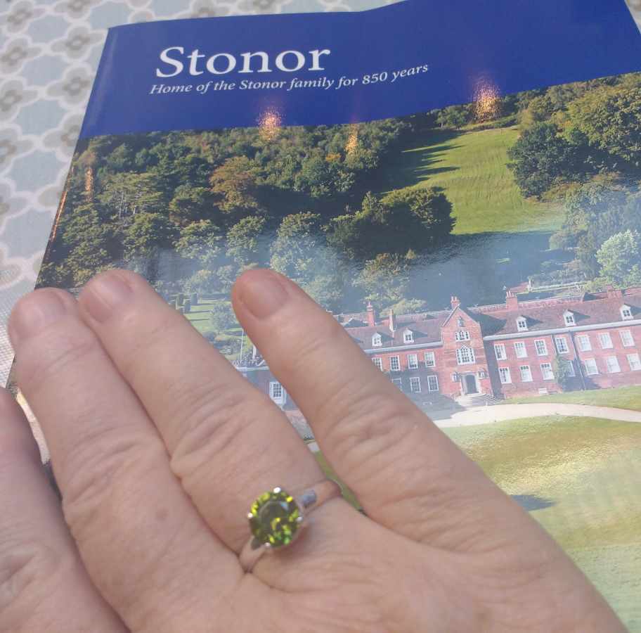 A hand wearing a green gemstone ring rests on a brochure titled Stonor featuring a large brick house and green grounds.
