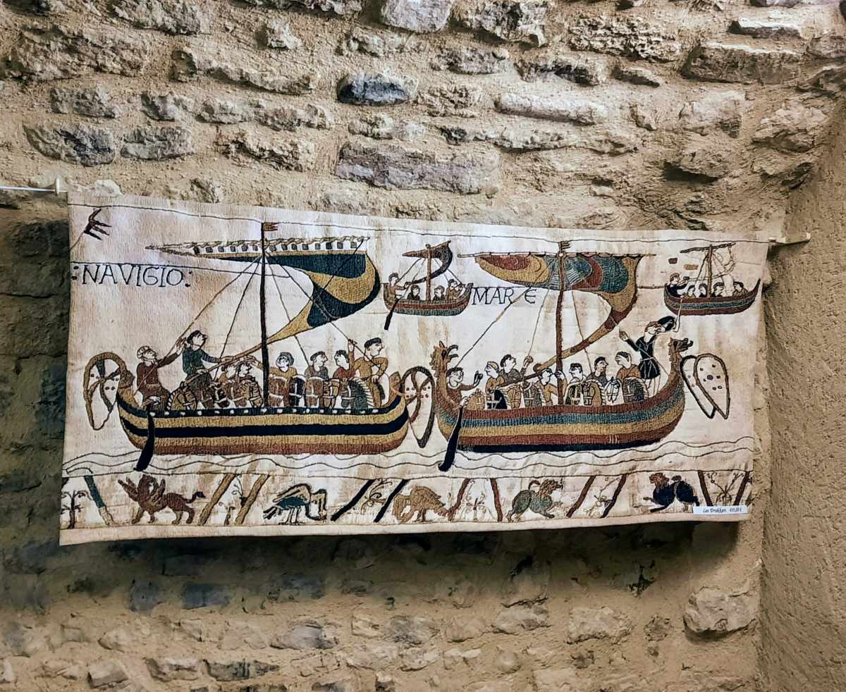 A tapestry depicting two ships with figures and the text NAVIGIO and MARE hangs on a stone wall.