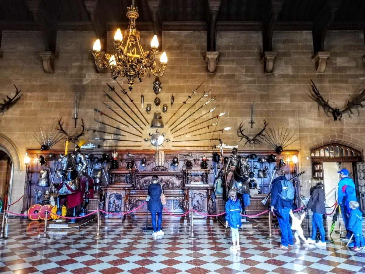People view a collection of antique armor and weapons displayed on a stone wall beneath a large chandelier.