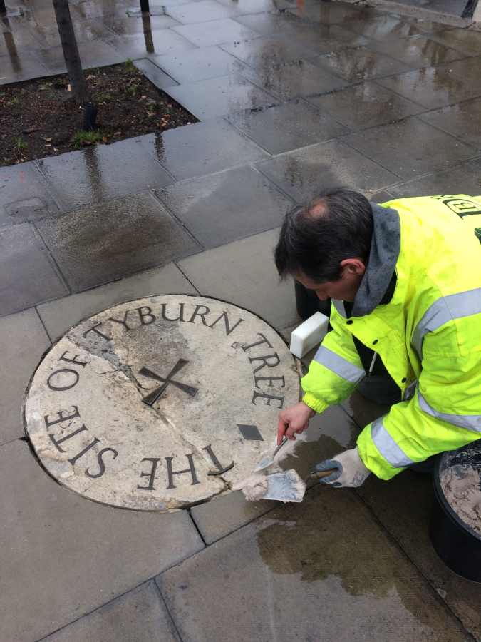 A person in a high-visibility jacket uses a trowel to apply mortar to a circular stone marker inscribed with text that says Tyburn Tree where the English were hanged
