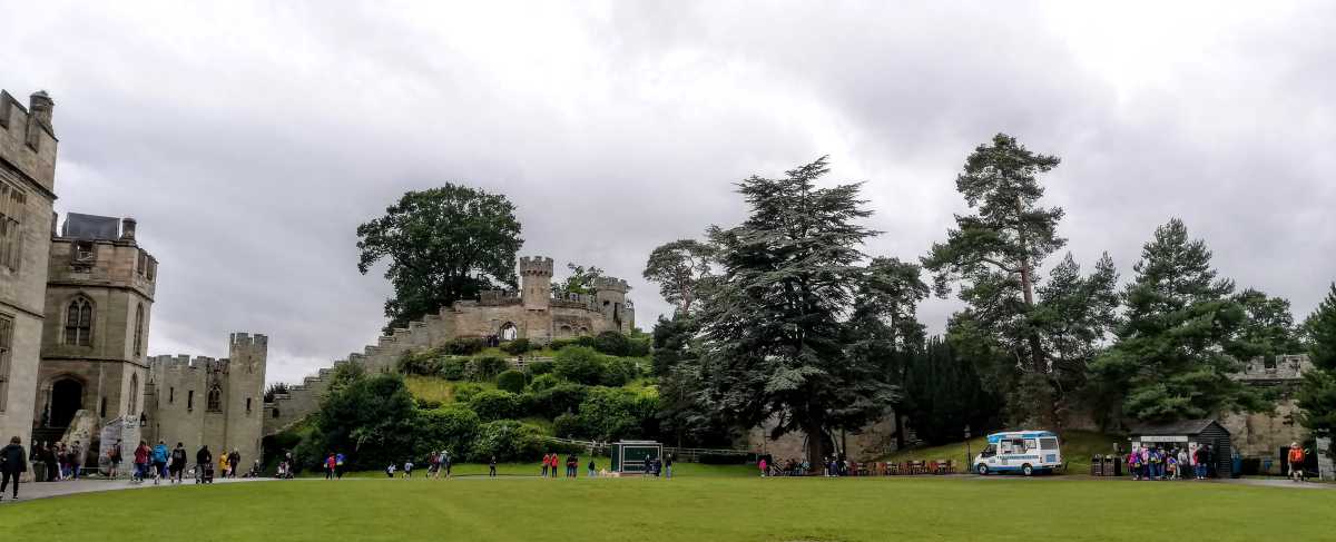 A stone castle with towers and walls sits on a grassy hill surrounded by trees under a cloudy sky.