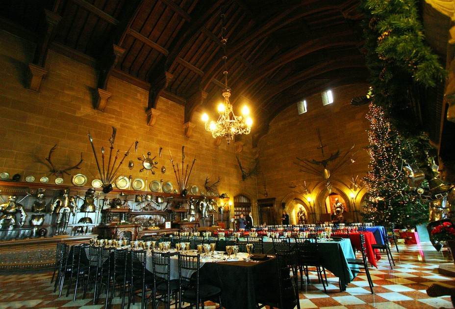 A large stone hall with a vaulted ceiling, a chandelier, long dining tables, and a decorated Christmas tree.