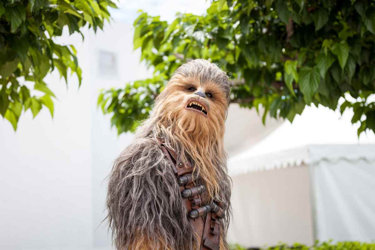 A person in a tall, furry, brown creature costume with a bandolier stands outdoors near green trees.