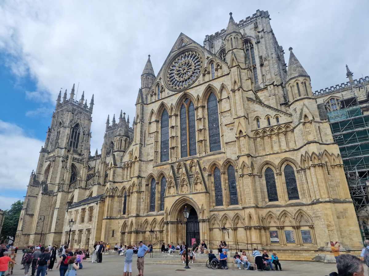 A large stone cathedral with a rose window and tall arched windows stands above a plaza with people sitting and walking.
