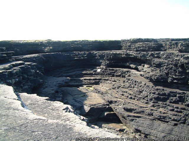 A natural, circular rock formation with layered, dark stone walls sits under a bright sky.