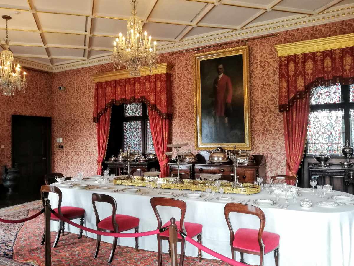 A formal dining room features a long table set for a meal, red patterned wallpaper, and a large portrait on the wall.
