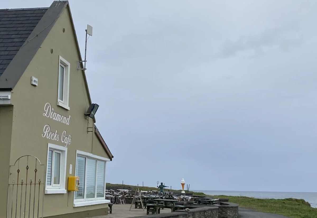 A green building labeled Diamond Rocks Cafe sits beside an outdoor patio with tables overlooking the ocean.