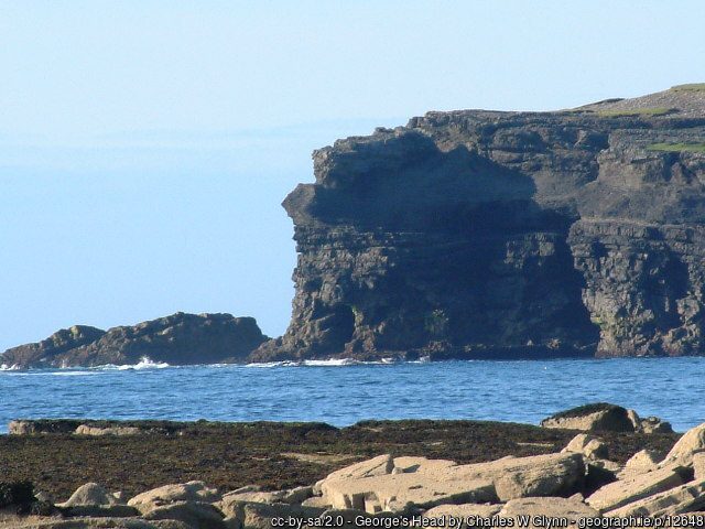 A rocky cliff face rises above the ocean, with a foreground of scattered stones and seaweed.
