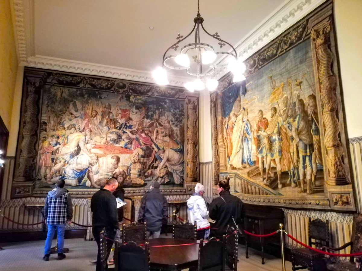 People stand in a room with large, ornate wall tapestries and a central chandelier.