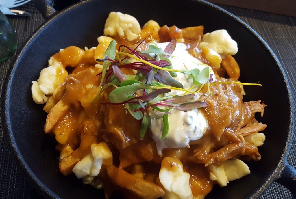 A Canadian national dish poutine. A bowl of poutine with the french fries and BBQ chicken topped with gravy and squeaky cheese curds topped with some sour cream 